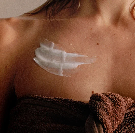 Person applying white cream to their shoulder with a brown towel draped over their shoulders.
